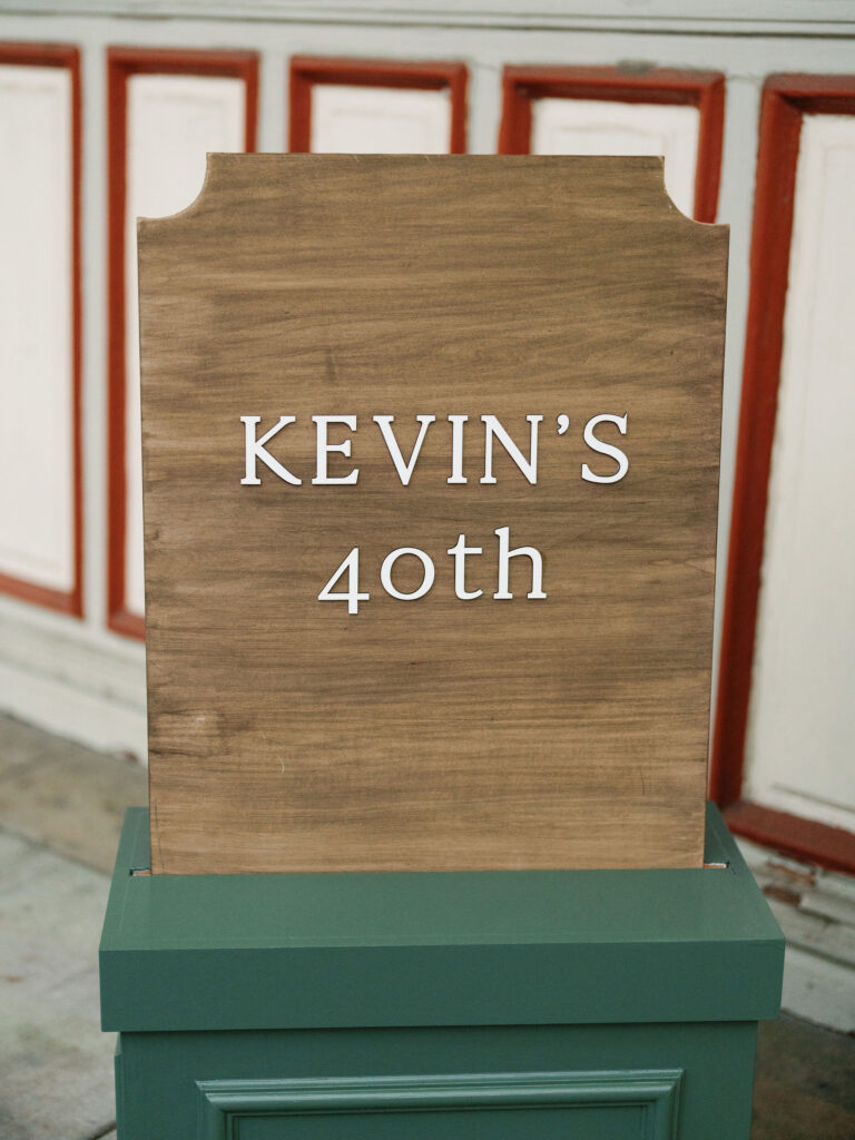 Wooden sign on a green stand reading "Kevin's 40th" in white letters, set against a backdrop of red and white paneling.