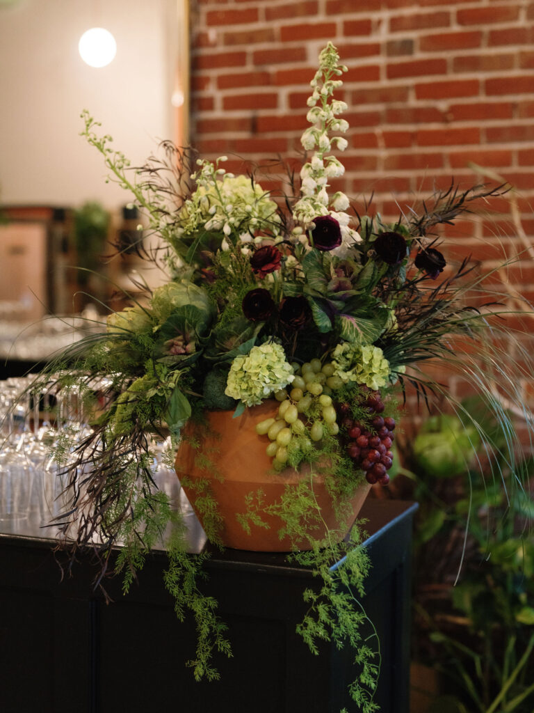 Floral arrangement with green and black flowers, leafy branches, and grape clusters in a terracotta pot, set on a dark surface against a brick wall backdrop.