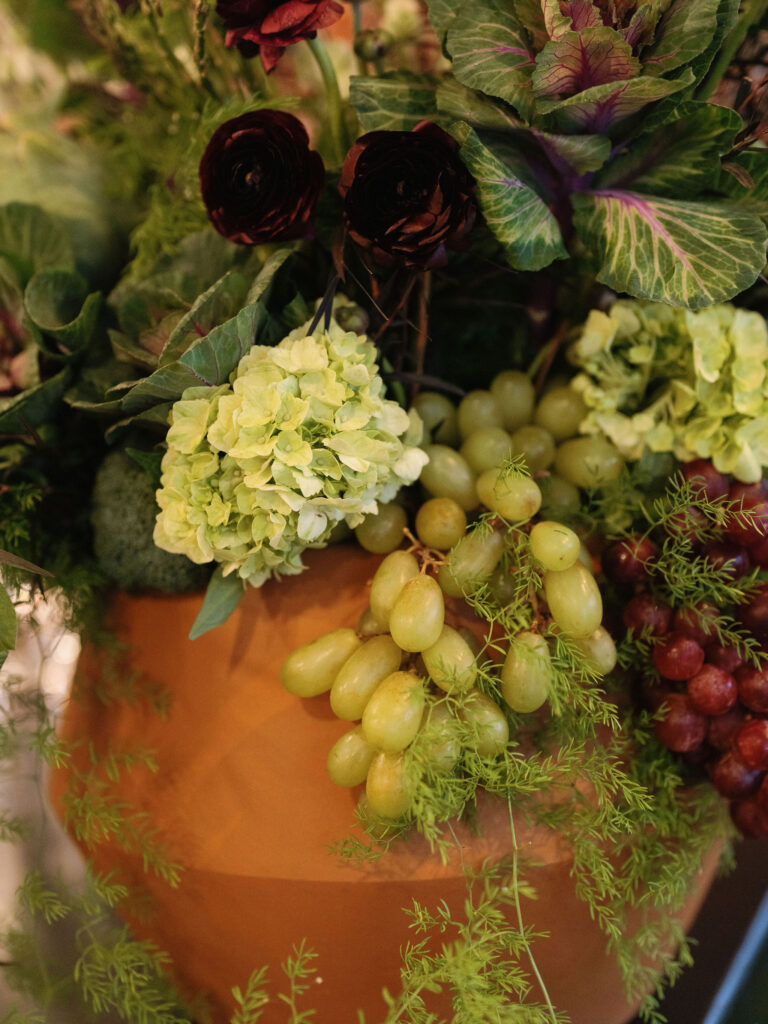 Arrangement of green grapes, red grapes, hydrangeas, and dark flowers with greenery in a large, terracotta planter.