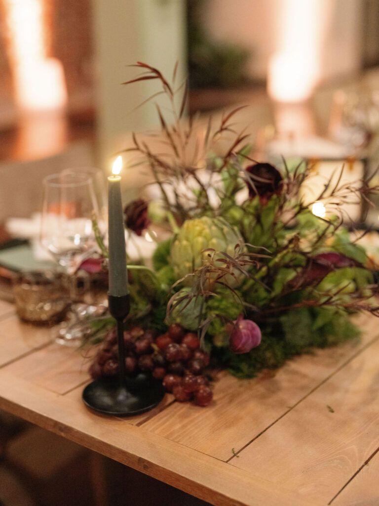A lit candle on a wooden table beside a floral centerpiece with greenery, purple accents, and grapes. Wine glasses and blurred dining setup in the background.