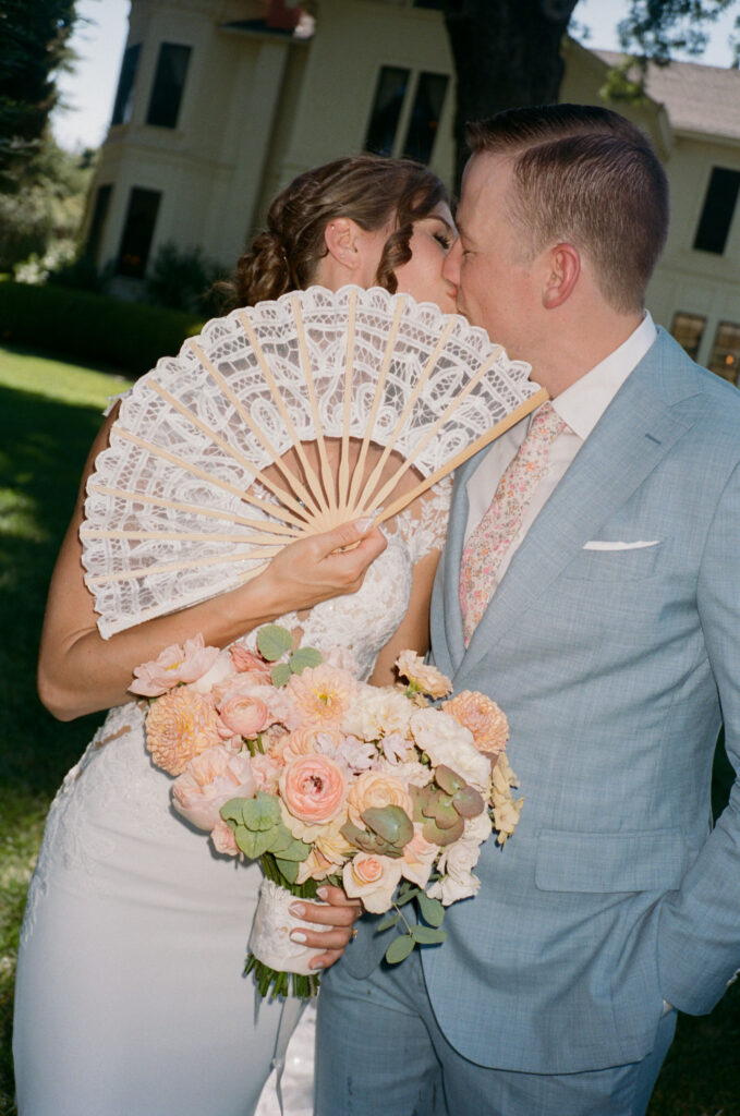 A bride and groom share a kiss beside a building. The bride holds a lace fan and a bouquet of flowers, while the groom wears a light blue suit.