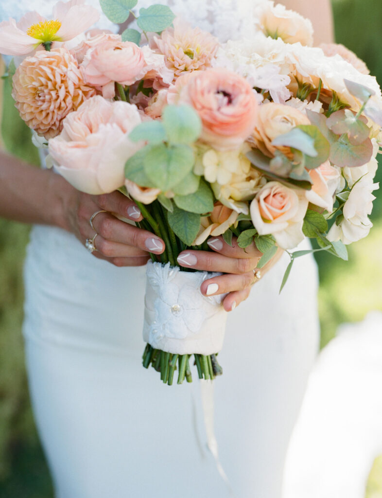 Bride holding a bouquet of peach and white roses, accented with greenery, wrapped in white lace.
