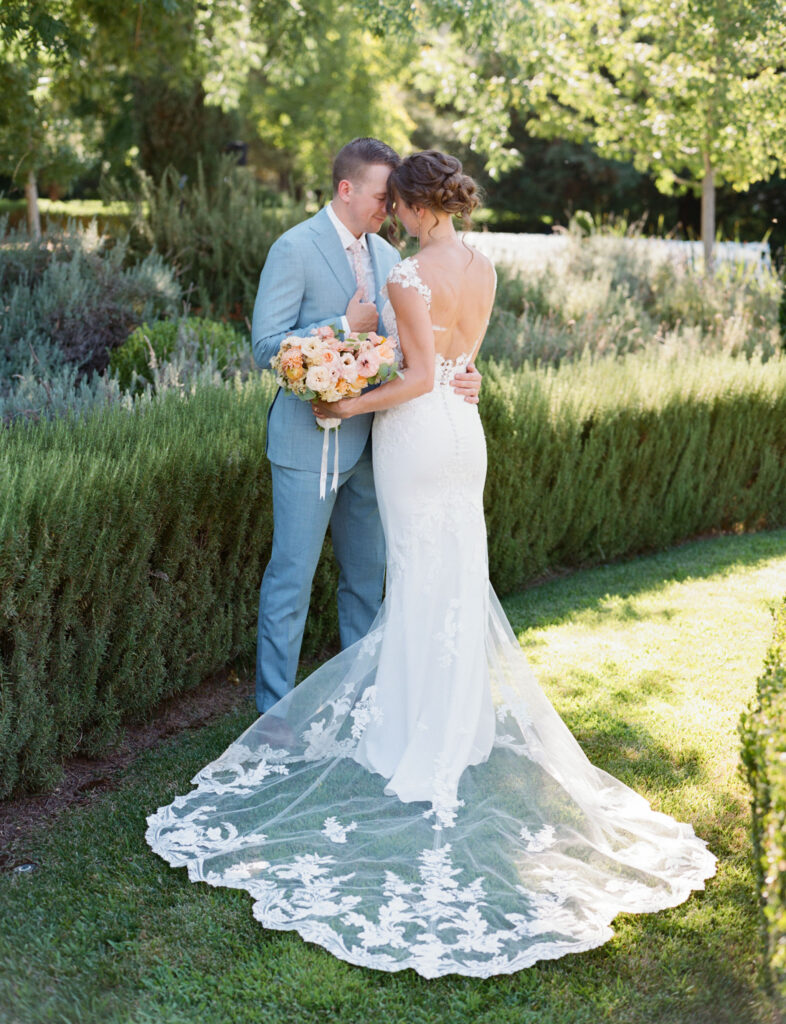 A bride in a lace dress and a groom in a light blue suit stand close in a garden, with the bride holding a bouquet of peach and white flowers.