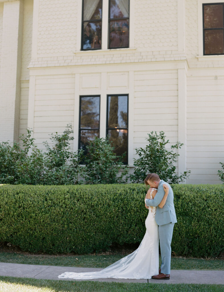 A couple in wedding attire embraces in front of a white building with large windows and a hedge.