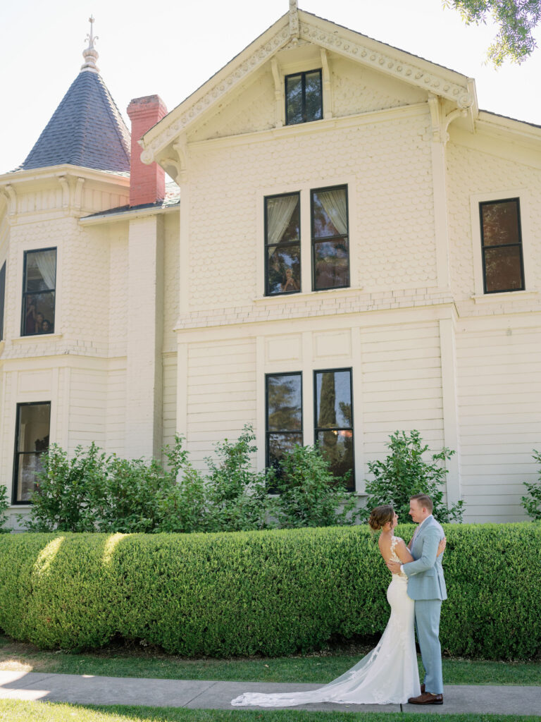 A bride and groom stand facing each other on a lawn in front of a large, light-colored house with tall windows and greenery.