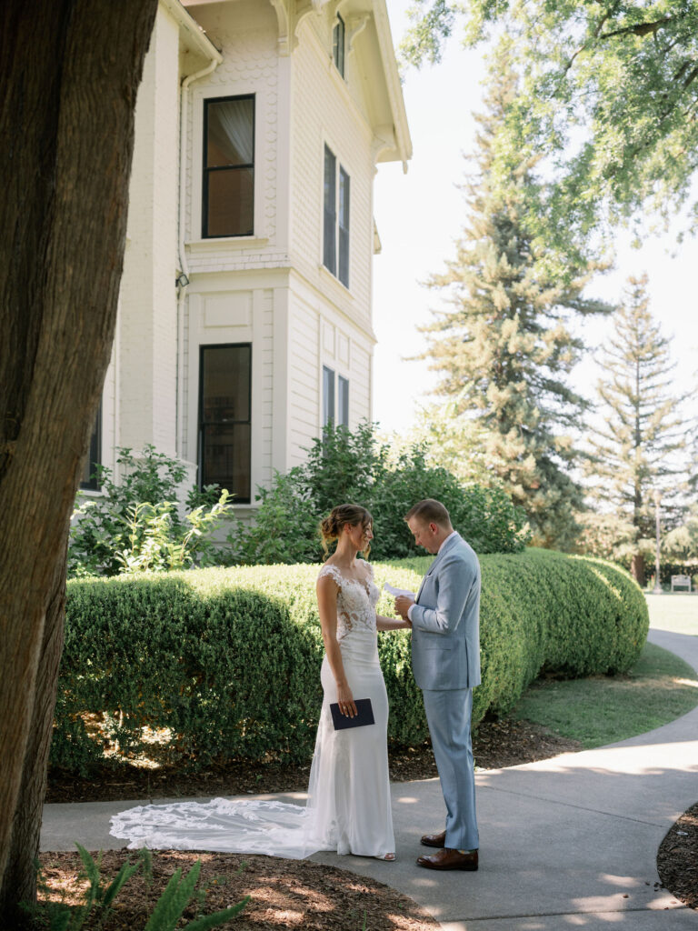 A couple stands together on a garden pathway beside a large house. Tall trees and hedges surround them. The woman wears a bridal dress, and the man is in a light blue suit.