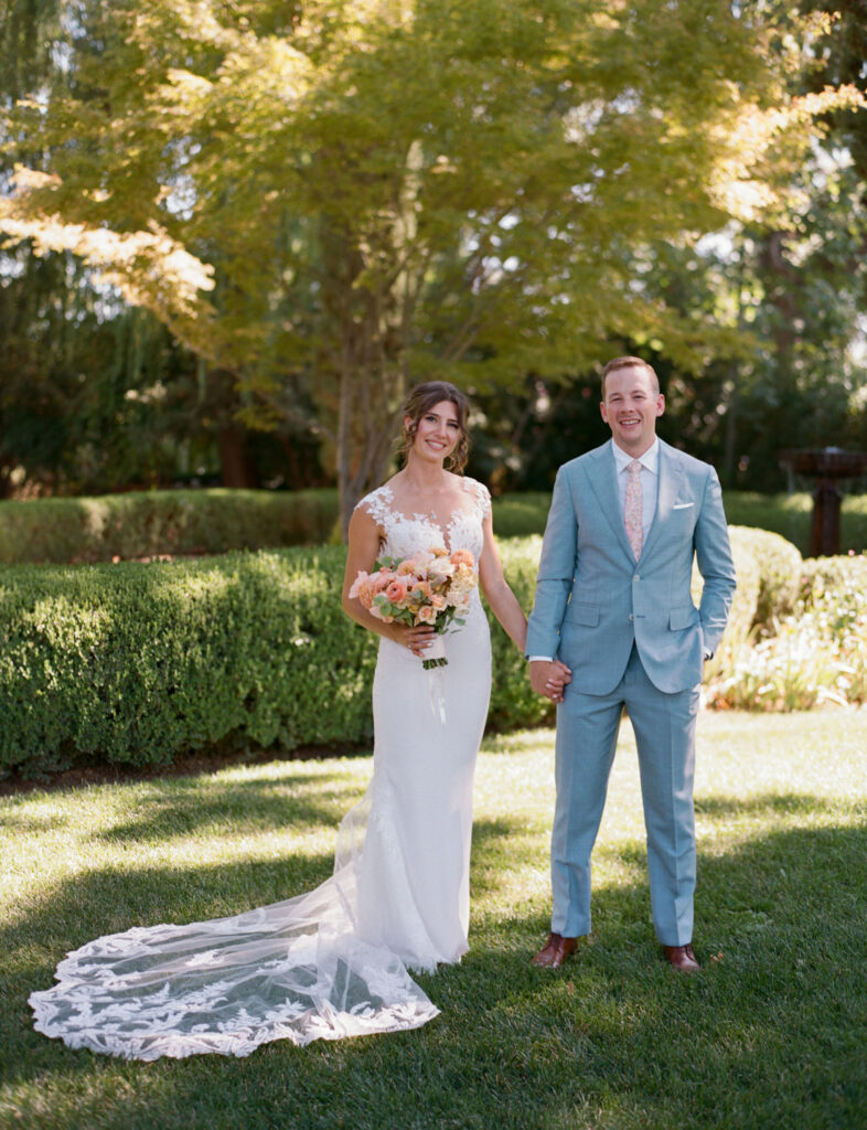 A bride and groom stand holding hands in a garden. The bride wears a white dress and holds a bouquet, while the groom wears a light blue suit. Greenery and trees surround them.