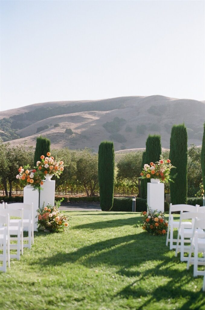 Outdoor wedding setup with rows of white chairs facing a floral altar. Tall cypress trees and rolling hills are in the background under a clear sky.