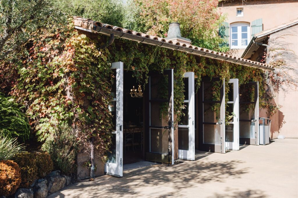 A building's exterior is covered in dense green and red ivy. Several open glass doors reveal an indoor area. The ground is paved, and there are shrubs on the left.