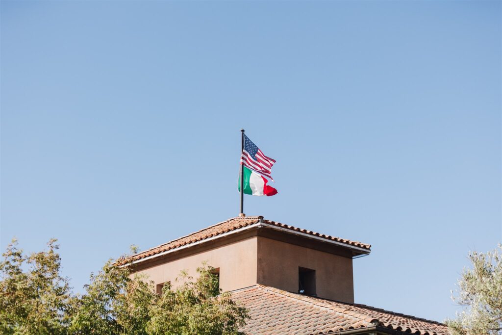 Two flags, American and Italian, on a tiled rooftop under a clear blue sky, surrounded by tree branches.