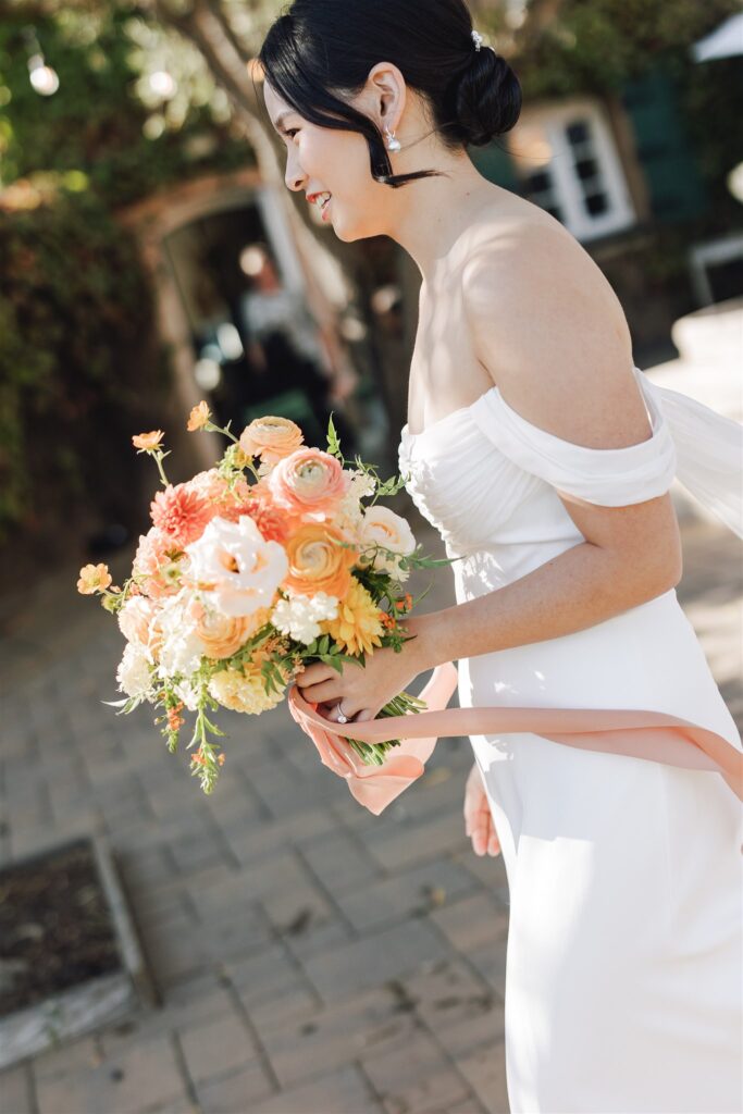 Bride in a white dress holds a vibrant bouquet of orange and white flowers, standing on a paved path with greenery in the background.