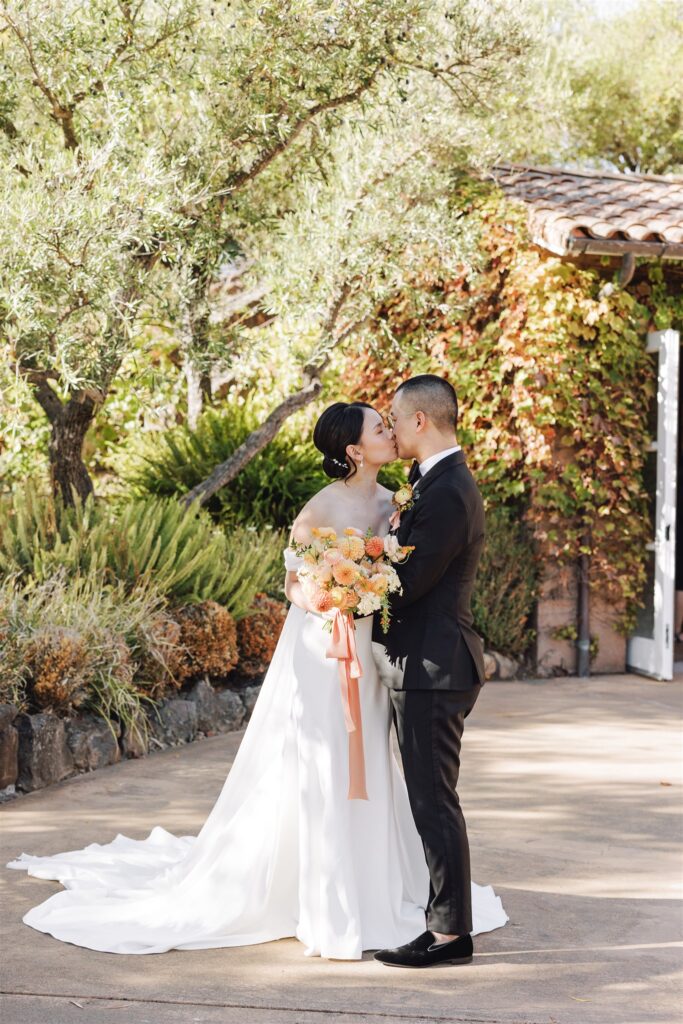Bride and groom sharing a kiss outdoors near a building covered in greenery. The bride holds a bouquet of yellow and peach flowers.