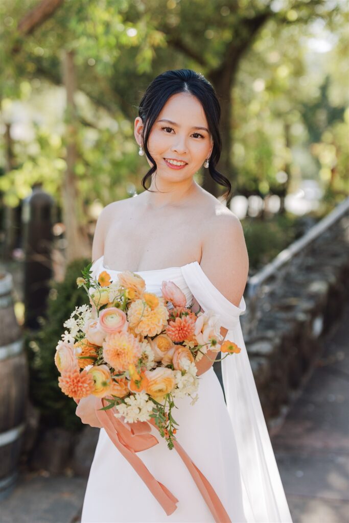 A bride in a white dress holds a bouquet of orange and cream flowers. She stands outdoors by a stone wall, with green trees in the background.