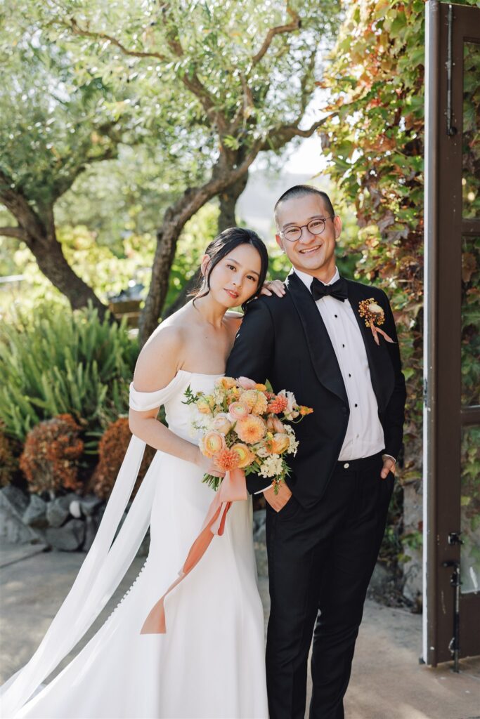 A bride and groom pose outdoors. The bride holds a bouquet of orange and yellow flowers, while the groom wears a black suit with a bow tie. They stand in front of trees and greenery.