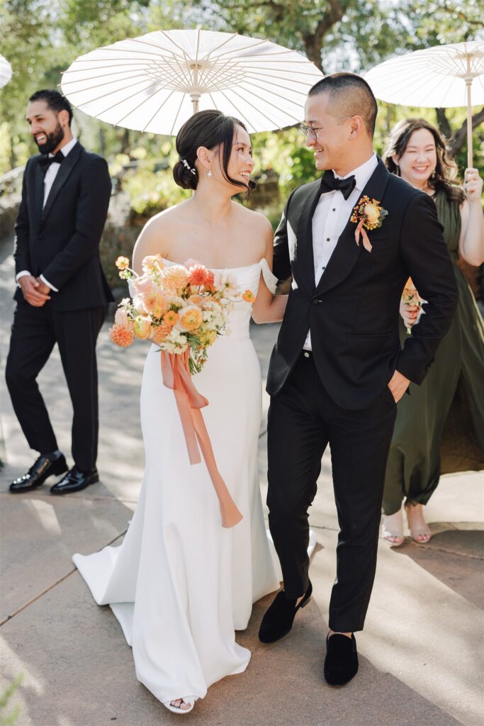 A bride and groom smile at each other while holding hands. The bride holds a bouquet, and people in formal attire stand around them with white parasols.