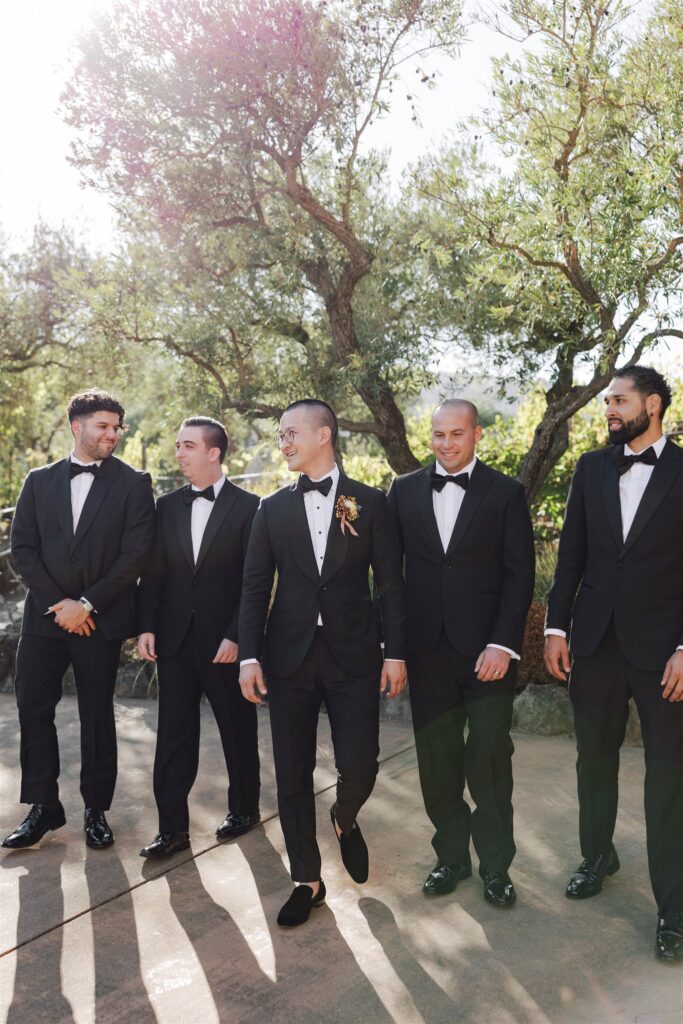 Five men in black tuxedos standing outdoors on a sunny day, with trees in the background. The central figure is looking left, while others face him, seeming engaged in conversation.