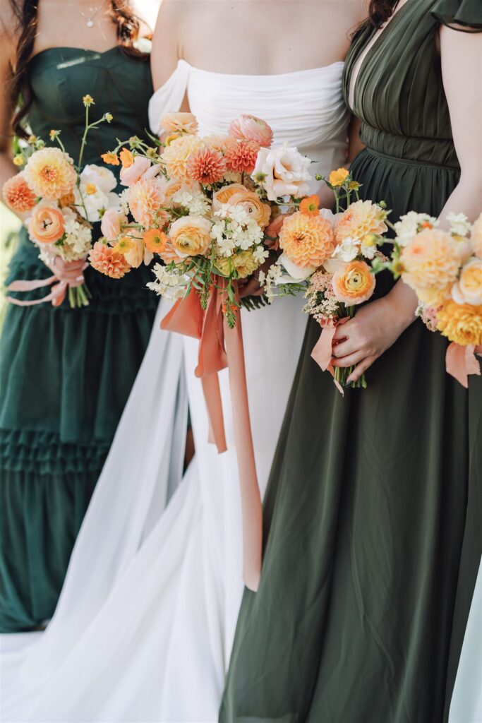 Three women in formal attire hold bouquets with orange and white flowers. The center woman wears a white dress, while the others wear dark green dresses.