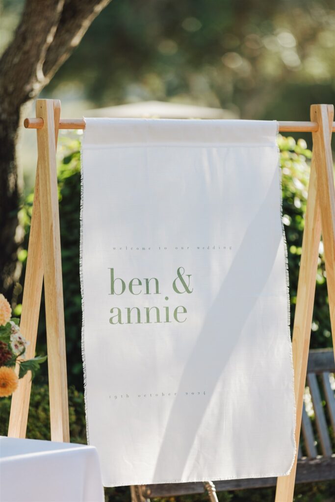White wedding banner on a wooden stand reads "welcome to our wedding, ben & annie, 8th october 2021" with flowers and greenery in the background.