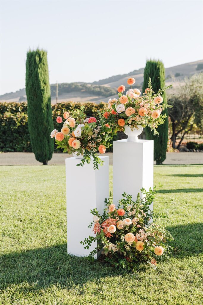 Floral arrangements with pink and orange flowers on white pedestals stand on a grassy area, with hills, tall trees, and hedges in the background.