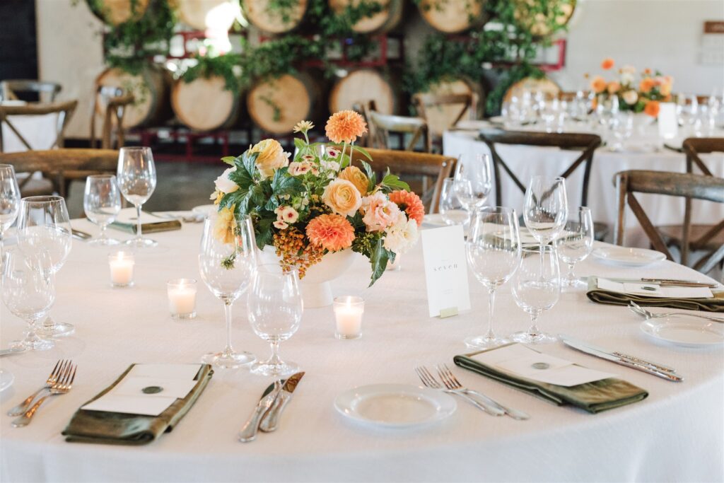 Elegant table setting with floral centerpiece, surrounded by wine barrels. Includes glasses, cutlery, and place settings on a white tablecloth with small candles.
