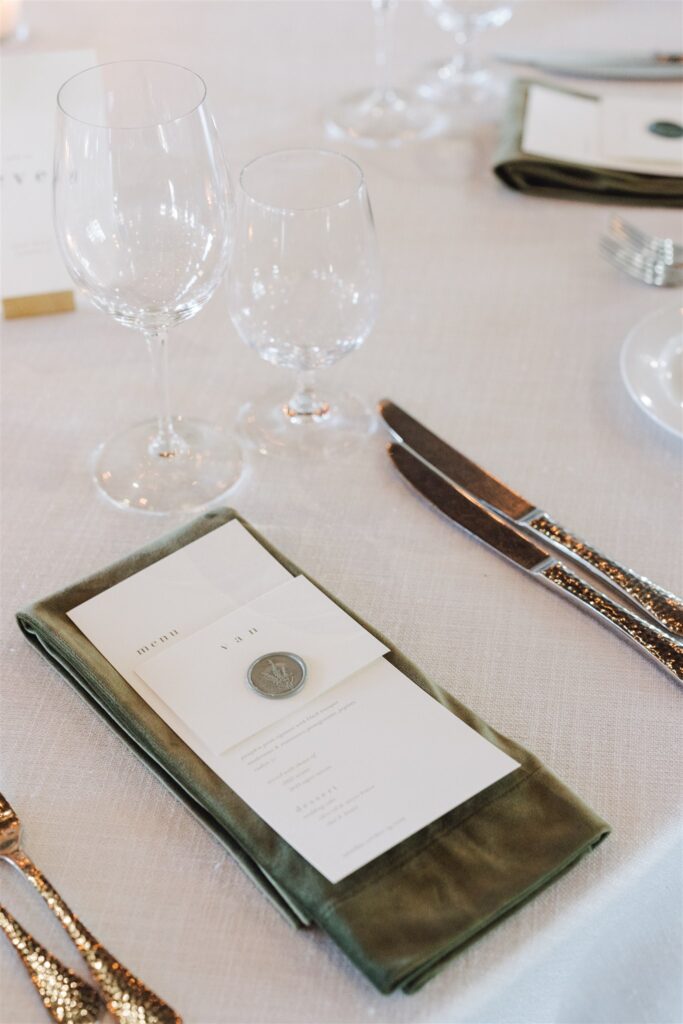 Table setting with two wine glasses, a fork, and knife beside a cloth napkin holding a menu card. White tablecloth background.