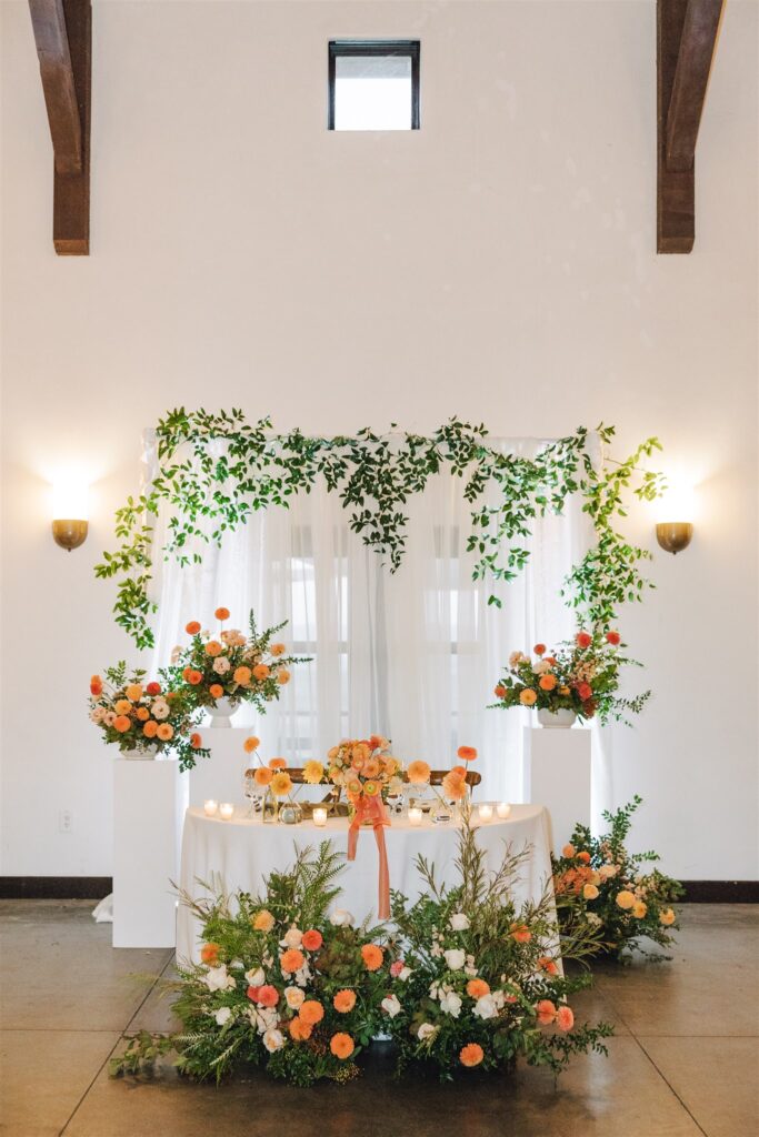 A decorated wedding table with white drapes, greenery, and orange and white floral arrangements. Candles are placed around the table in a well-lit room.