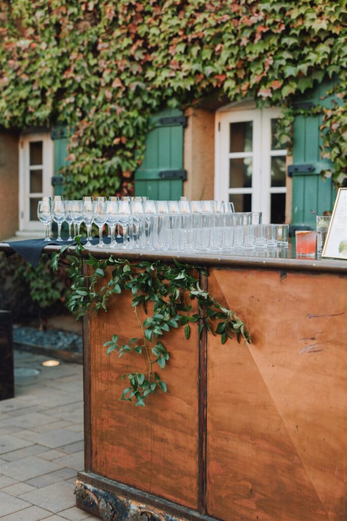Outdoor bar setup with empty wine and champagne glasses on a wooden counter. Green ivy creeps up the wall behind, with windows and green shutters in view.