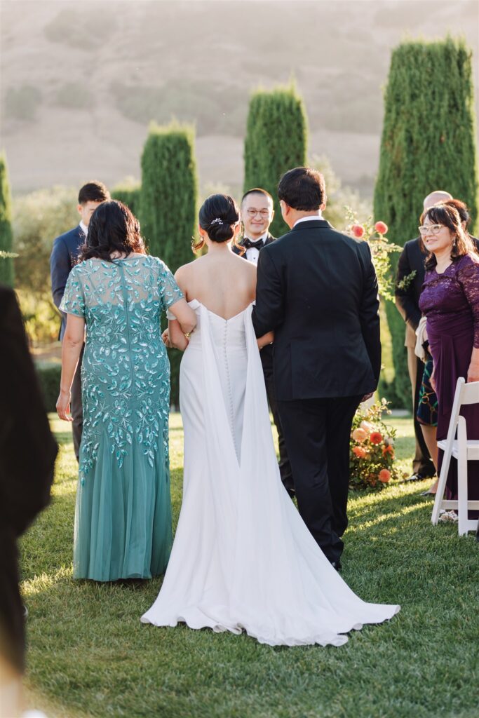 A bride in a white dress is walking down the aisle with two people, one on each side. Trees and flowers are in the background.