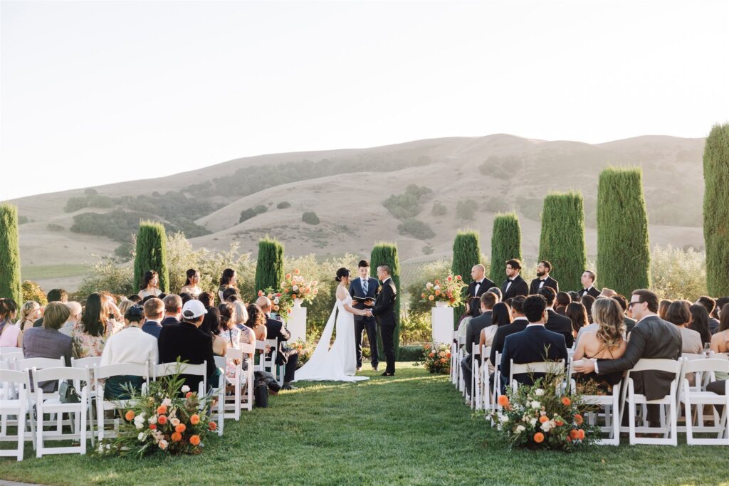 Outdoor wedding ceremony with a couple at the altar, surrounded by bridesmaids and groomsmen. Guests seated in rows on a lawn amidst floral arrangements. Hills and clear sky in the background.