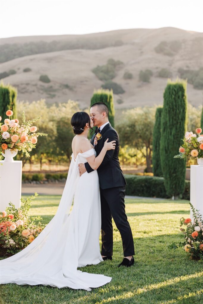 A couple embraces outdoors in formal attire, surrounded by floral arrangements and greenery, with hills in the background.