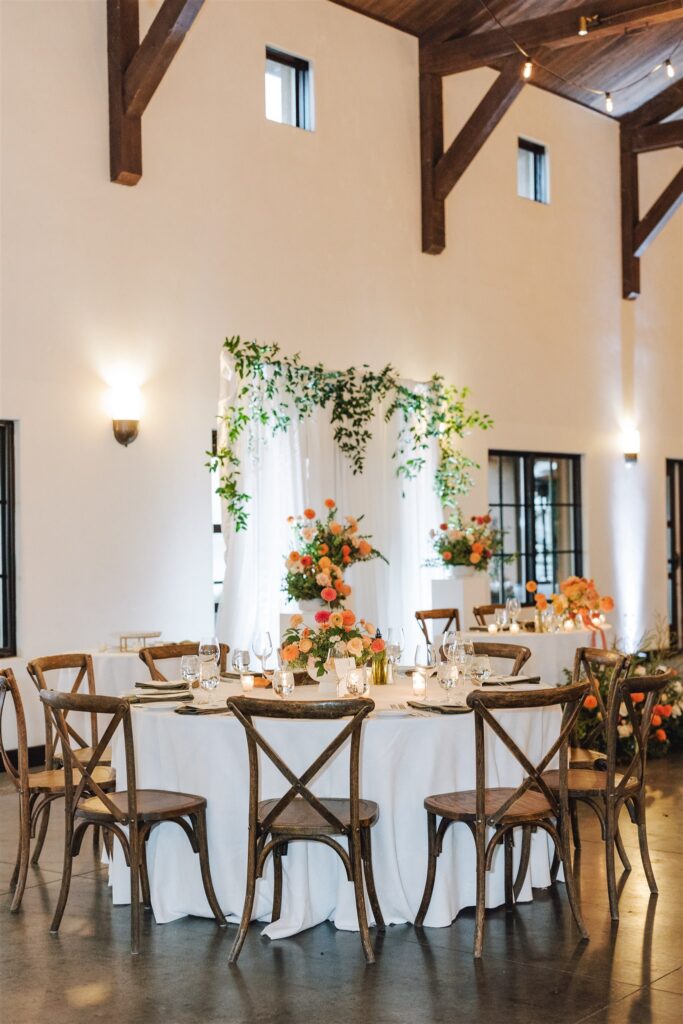Elegant dining setup in a warmly lit room, featuring round tables with white tablecloths, wooden chairs, and vibrant floral arrangements. An arch with greenery and flowers is in the background.