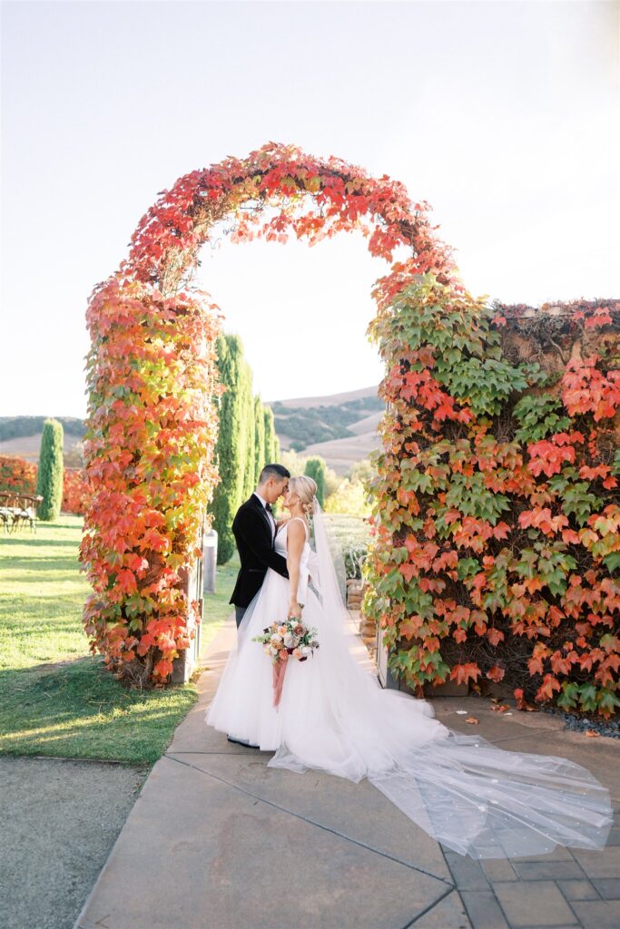 A bride and groom stand facing each other under an archway covered in vibrant autumn leaves, with a scenic backdrop of grass and hills. The bride holds a bouquet, and her veil flows behind her.
