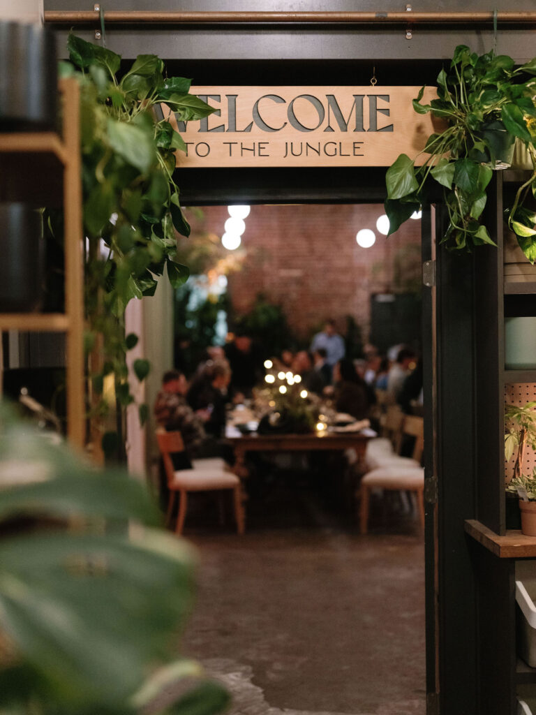 Entrance to a dining area with plants, featuring a sign saying "Welcome to the Jungle." People are seated at a table in the background.