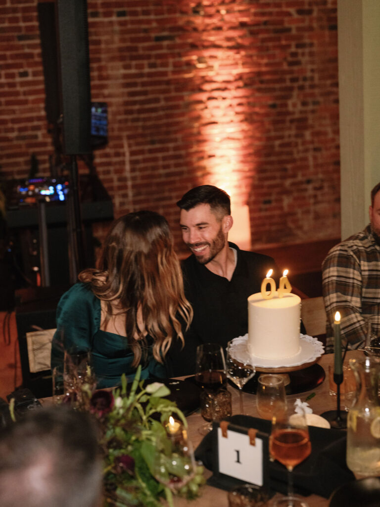 A couple sits at a table with a white cake topped with number-shaped candles. The setting is a brick-walled room with dining decorations and drinks.