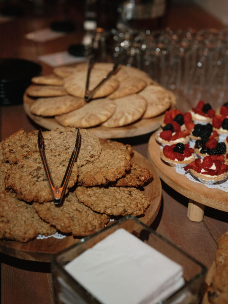 Assorted cookies, pastries, and berry-topped desserts on wooden platters with tongs, beside a stack of napkins on a wooden table.