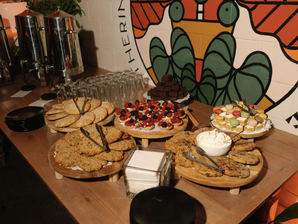 A dessert buffet with assorted cookies, pastries, macarons, brownies, and a bowl of cream on wooden stands. Silverware and stacked glasses are also on the table.