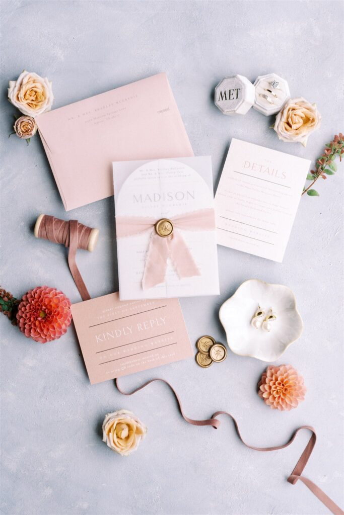 Flat lay of elegant wedding stationery with pink and beige cards, roses, a spool of thread, a ribbon, and decorative elements on a light textured background.