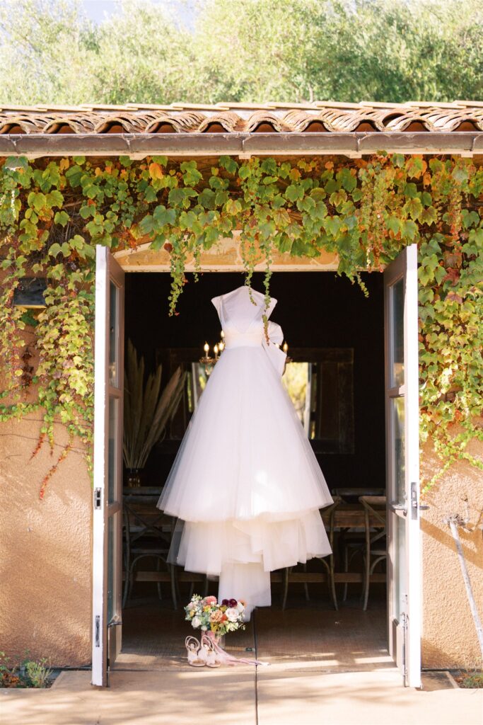 A wedding dress hangs in a doorframe draped with greenery, above a pair of shoes and a bouquet on the ground.