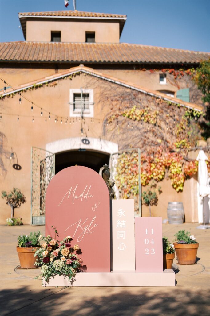 Wedding venue entrance with terra cotta building. Sign reading "Maddie & Kyle" with date 11/04/23, beside floral arrangement and string lights.