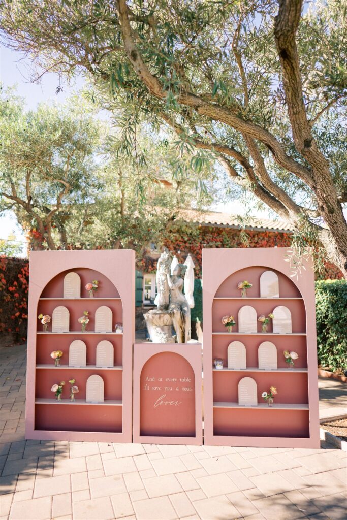 A pink seating chart display with shelves is adorned with small flower arrangements and plaques. A garden statue is visible behind, framed by leafy trees under a clear blue sky.