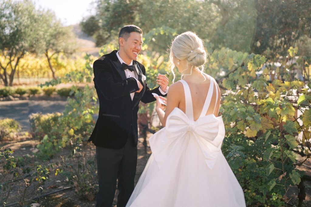 A couple stands outdoors. The man wears a black suit, and the woman wears a white dress with a large bow on the back. They face each other, surrounded by greenery.