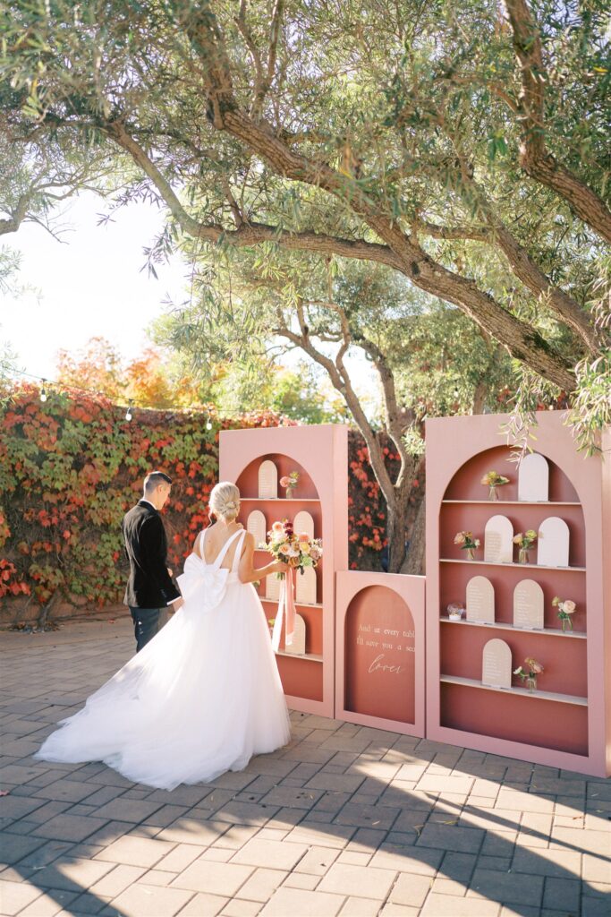 A bride and groom stand in front of a pink decorative display with arches, containing flowers and placards. They are outside, with trees and a vine-covered wall in the background.