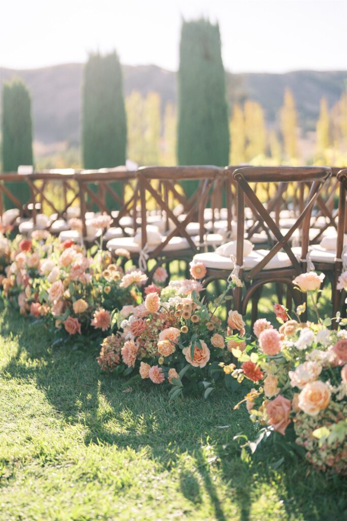 Outdoor wedding ceremony setup with wooden chairs arranged in rows. The aisle is lined with abundant peach and pink floral arrangements. Bushy trees and hills are visible in the background.