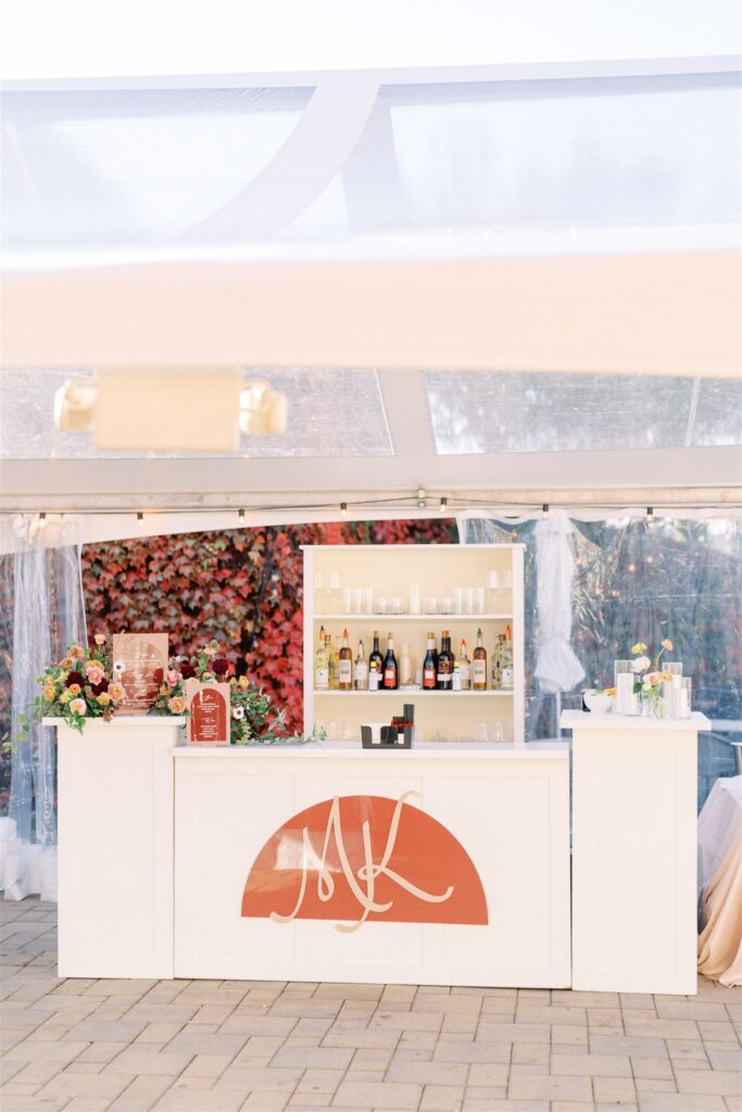 Outdoor bar setup under a transparent tent with a variety of drinks and glasses; decorated with flowers and featuring a monogram design on the front.