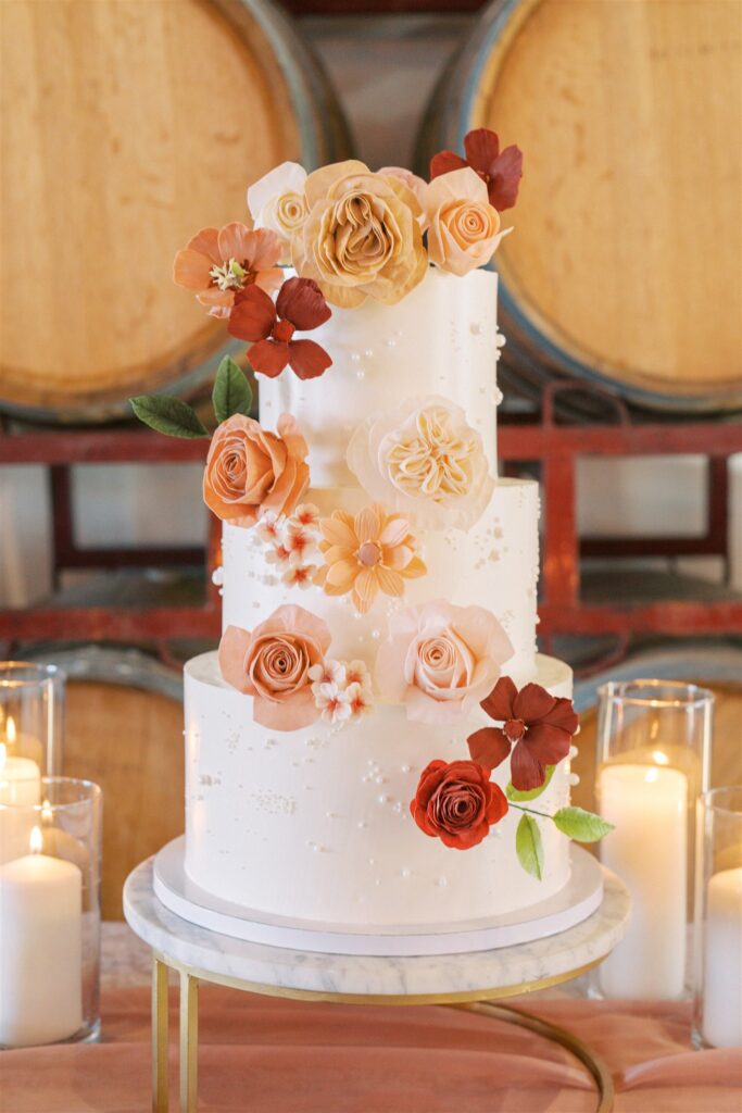 Three-tiered white cake adorned with pink and orange flowers, surrounded by lit candles on a marble stand, set against wine barrels.