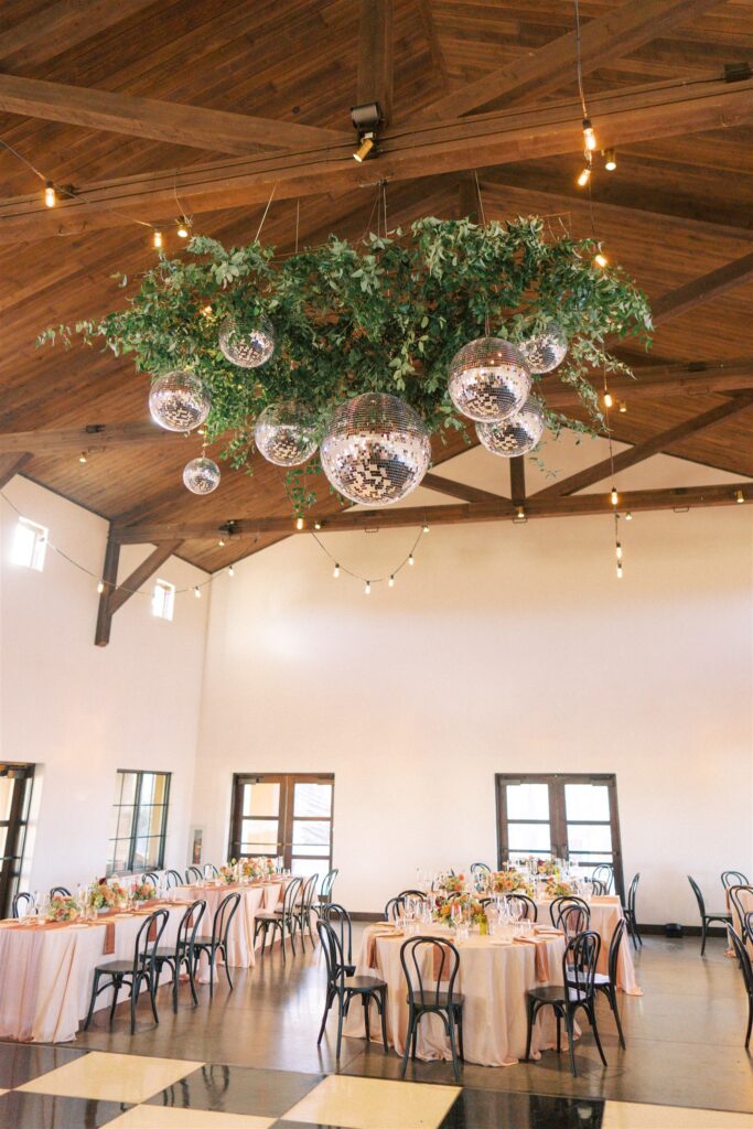 Spacious banquet hall with wooden ceiling, tables set for dining, black chairs, pink tablecloths, and a large hanging decoration of greenery and reflective spheres.