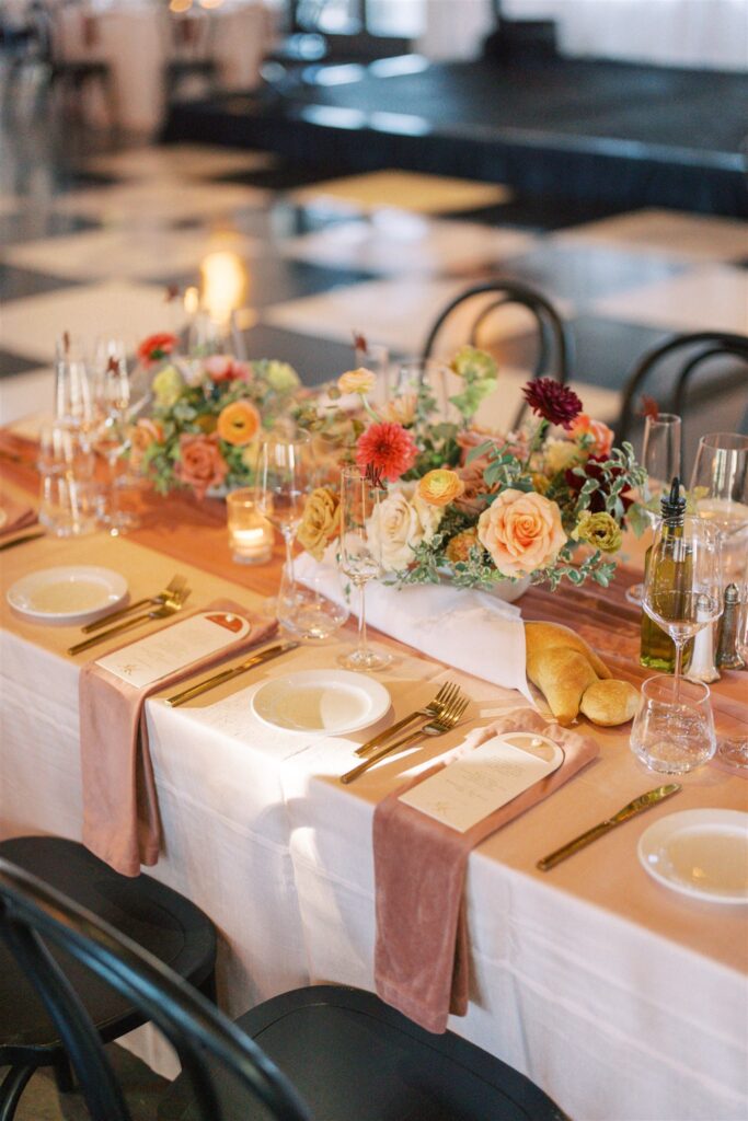Elegant table setting with floral centerpiece, wine glasses, candles, plates, cutlery, and a baguette on a white tablecloth. Checkerboard floor visible in the background.