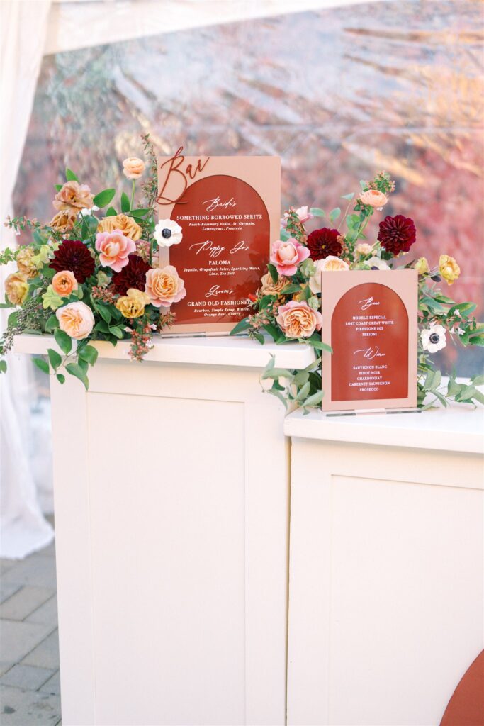Wedding bar setup with white counter adorned with colorful flowers. Two red signs display drink options and names.