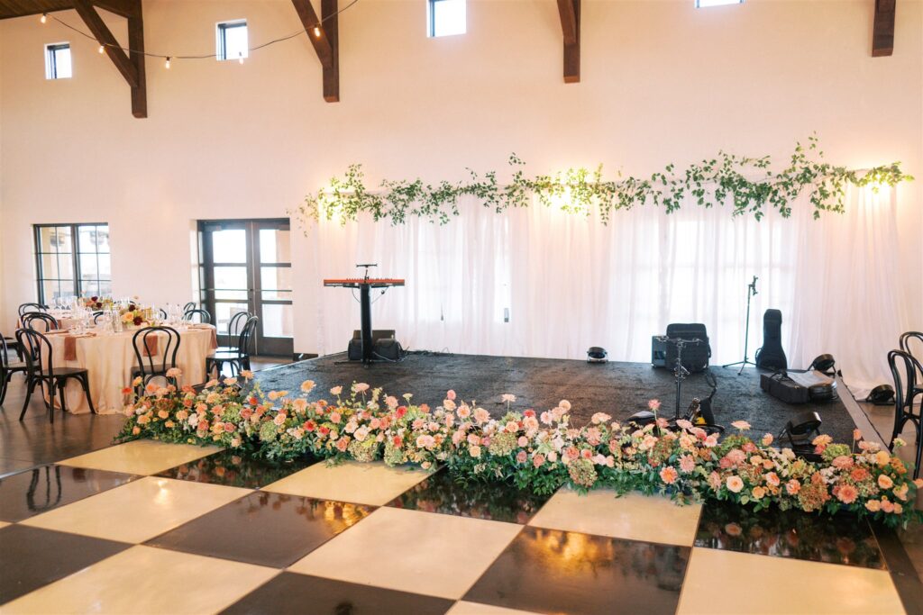A decorated event stage with musical instruments and floral arrangements on the floor. A checkerboard dance floor and dining tables are in the foreground.