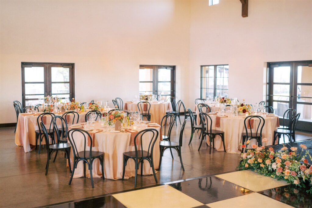 A spacious event room with round tables set for a gathering, featuring black chairs and floral centerpieces. A black-and-white checkered floor is in the foreground.